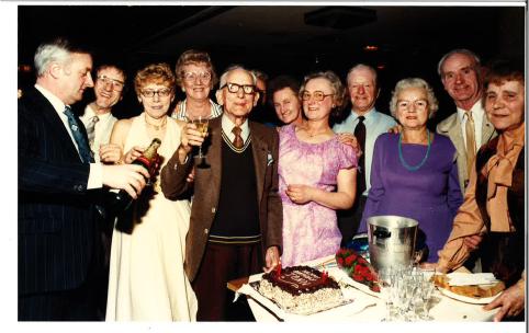 A colour photo of a crowd celebrating, with Mr John Lamb at the centre, raising a glass. On the table in front of him is a cake, with white and brown icing and red candles.