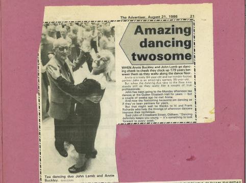 A scan of a newspaper clipping with the headline “Amazing dancing twosome”, with an image to the left of John dancing with Annie Buckley in a hall. The hall has a wooden floor, and many other couples dancing in the background, who are blurred.