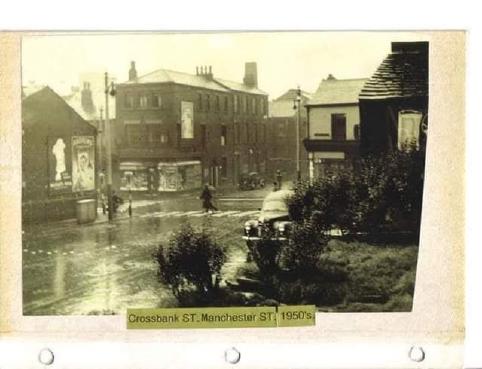 A scan of a yellowed photograph of a road junction at Crossbank Street. The road pictured is wet with rain, and there is a black car in the foreground behind some shrubbery. There is a shop on the corner, and some billboards opposite.