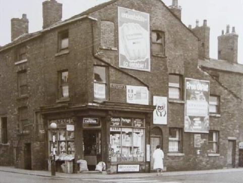 A sepia tone photo of a corner shop, in a three storey brick building, with lots of advertisements and billboards all over the side facing the camera. There are a couple of seemingly broken windows on the top floors, and a woman in a white knee length coat stood in front of the shop.