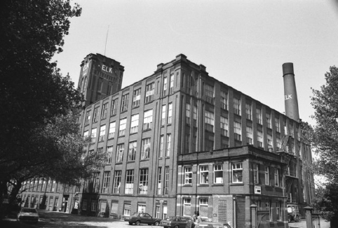 A black and white photograph showing Elk Mill, a five storey brick building with lots of windows, and a smokestack to the rear which reads ELK in large white letters.