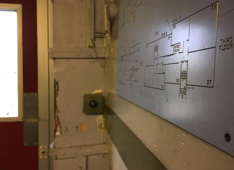 An image of a corner of a hallway area. In the foreground is a large plastic map showing the layout of the tower. Also visible is a ‘push to exit’ button, and the side of a red door with a window in, showing daylight outside.