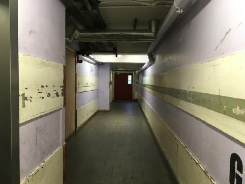 A photo looking down a corridor, which is dimply lit. There are panels running down each side of the corridor, painted a light yellow with an olive green stripe. The floor is a grey tile. Everything in the image is visibly dirty, with marks all over the floor and walls.