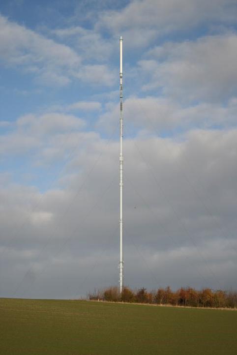 A colour photo of a white TV mast against a cloudy sky. The mast is very tall, with lots of cables tensioned between it and the ground. The mast is in a field, with some trees at the base.