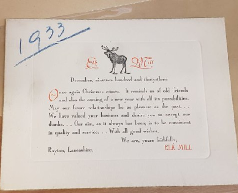 A photo of a white card on brown paper. At the top left of the card ‘1933’ is written in blue crayon. On the card is an illustration of an elk, surrounded by ‘Elk Mill’ in decorative red lettering. The card reads ‘December, nineteen hundred and thirty-three. Once again Christmas comes. It reminds me of old friends and also the coming of a new year with all its possibilities. May our future relationships be as pleasant as the past… We have valued your business and desire you to accept our thanks… Our aim, as it always has been, is to be consistent in quality and service… With all good wishes, We are yours faithfully, ELK MILL. Royton, Lancashire’