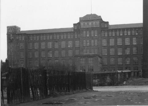 A black and white photo of Summervale Mill, a five storey brick building with a sign at the top which reads S DODD & SONS Ltd. There are large windows throughout the building. There is a rickety wooden fence in the foreground of the image.