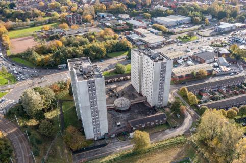 A colour aerial photo of the two towers, which are at the centre of the frame. You can see the space between the towers, with a structure connecting them on the ground floor. There are terraced houses to the right of the towers, and roads all around and behind them.