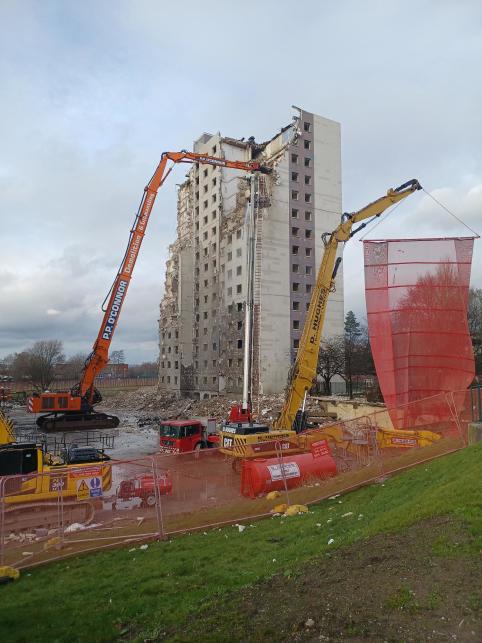 A colour photo of the towers during demolition. In the foreground, there is industrial fencing and a red net being lifted to control debris, with demolition in progress on the tower in the background. Only one of the towers is in frame, and over half of it is rubble.