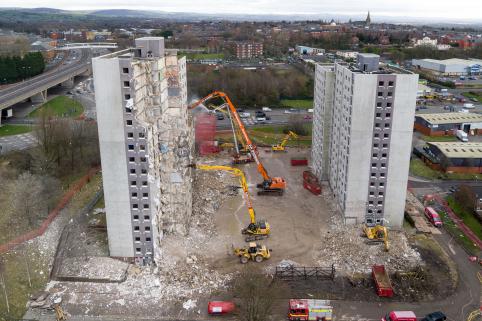 An aerial colour photo of the towers, with multiple machines working on demolishing them. The tower to the left of the image has been partially demolished, with only its left side still standing.