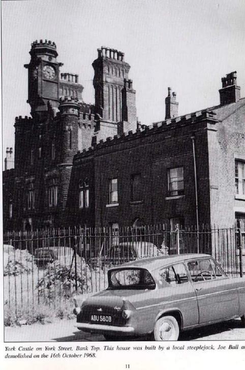 A black and white image seemingly copied from a publication or newspaper, which features a row of houses with one house featuring a number of chimneys, turrets, and other castle-like features. It sticks out from the row of houses a long way. There is an iron fence and some cars parked in the foreground of the image. At the bottom of the image there is a caption which reads “York Castle on York Street, Bank Top. This house was built by a local steeplejack, Joe Ball and demolished on the 16th October 1968”